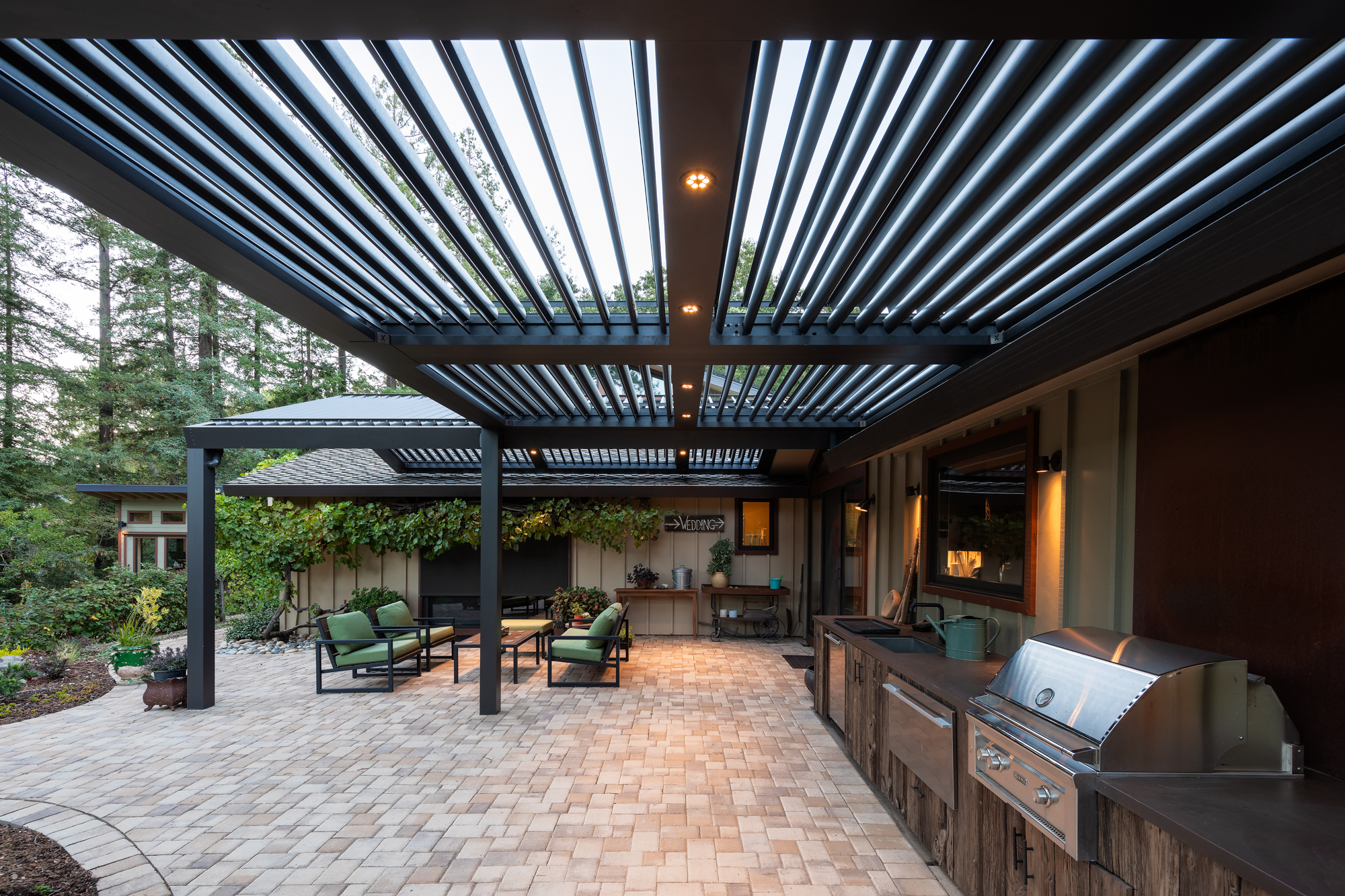 Charcoal motorized pergola over an outdoor kitchen and lounge area with redwood backdrop.