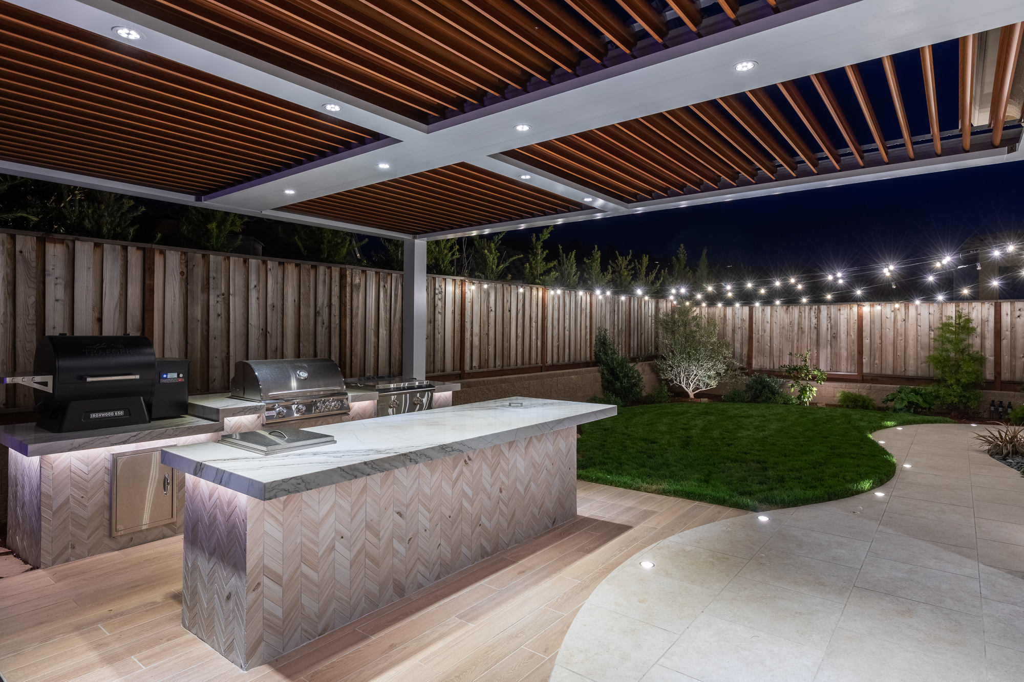 Integrated LED downlights inside a StruXure motorized pergola over an outdoor kitchen at night.