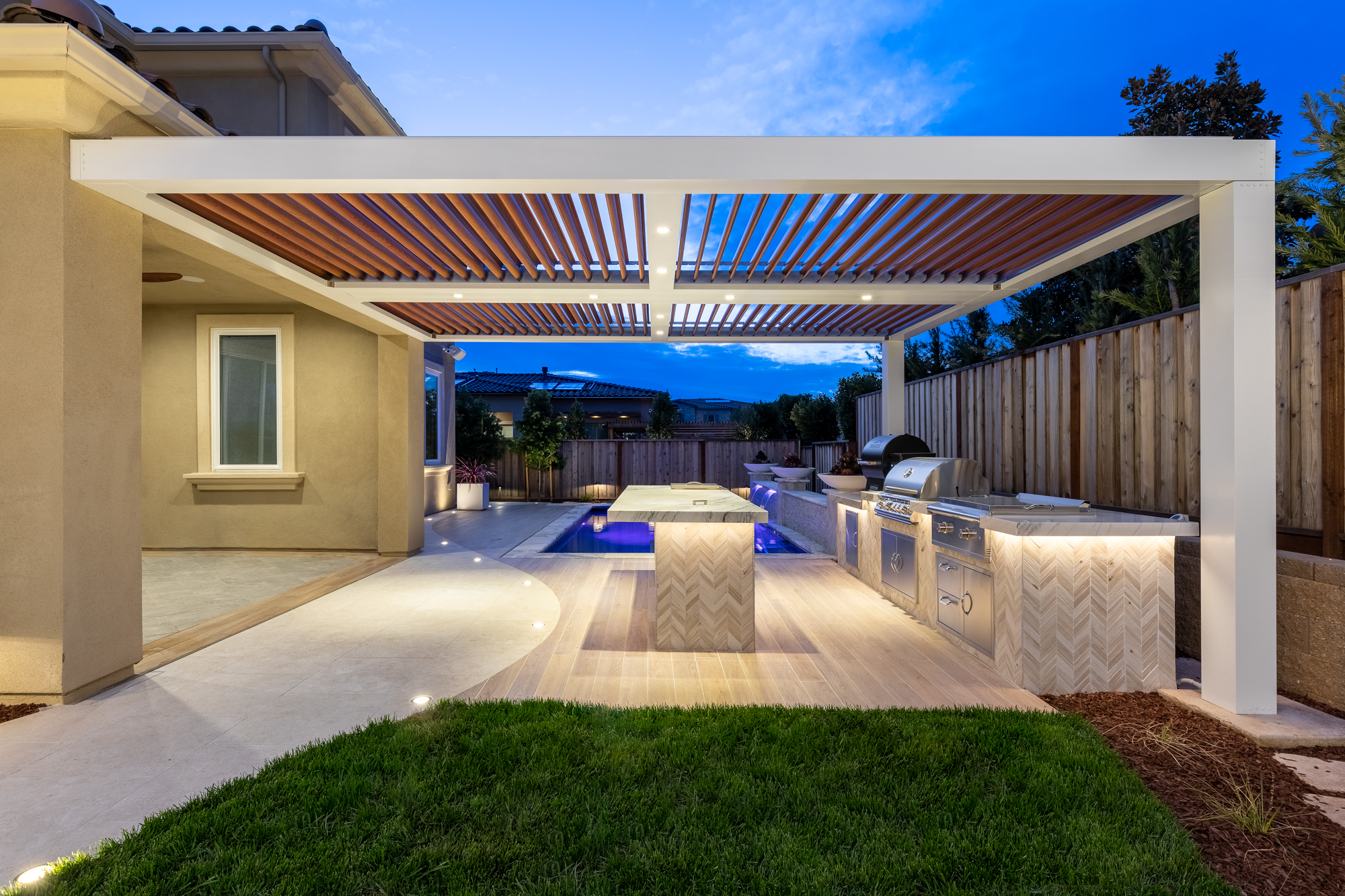 White pergola with wood-tone louvers over an outdoor kitchen and pool at twilight.