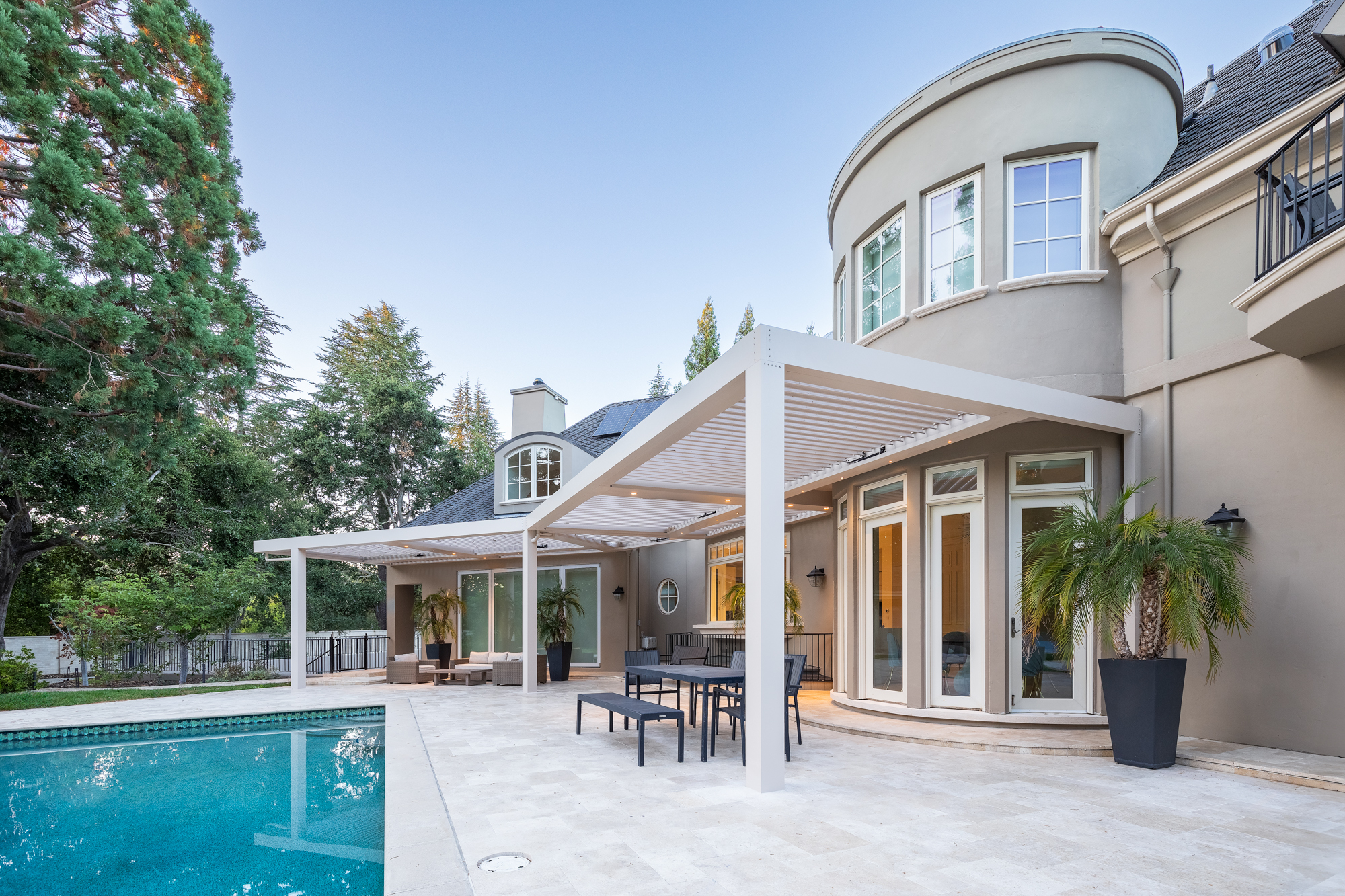 White motorized louvered pergola spanning a luxury poolside patio at dusk.