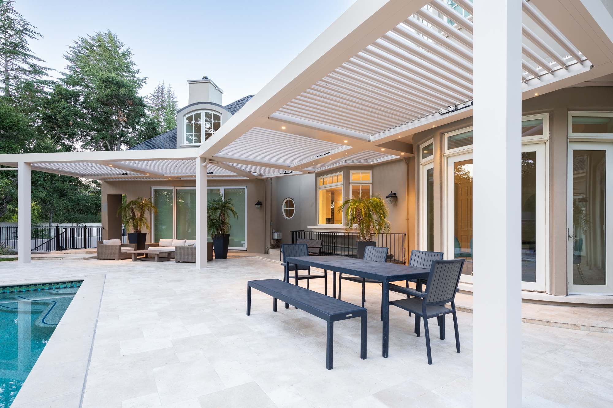 Outdoor dining set under a motorized louvered pergola next to a swimming pool.